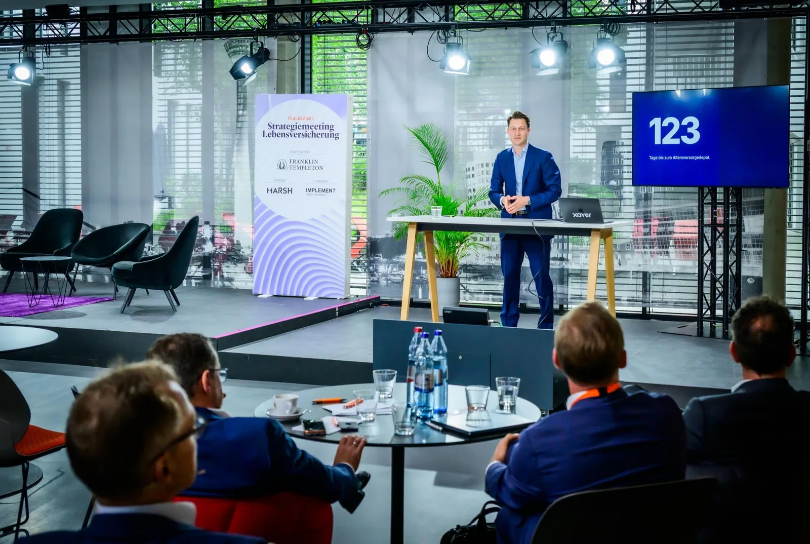 Max Bachem am Stehtisch auf der Bühne des Handelsblatt Strategiemeetings Lebensversicherung, auf dem Bildschirm „123 Tage bis zum Altersvorsorgedepot“.